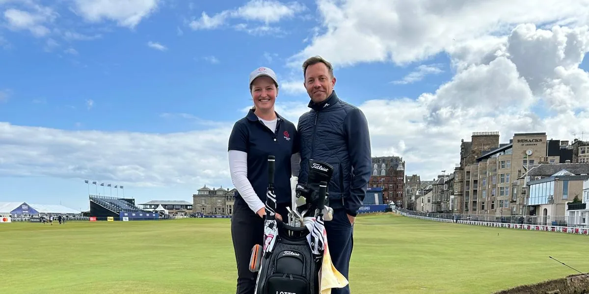 A coach and a player with a tour bag on the Swilcan Bridge at St Andrews, the Old Course behind them.